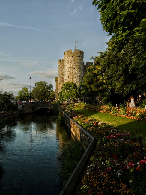 The image depicts a scenic view of Hertford Castle surrounded by vibrant flower beds with a variety of colorful blooms, including pink, orange, and yellow flowers, neatly arranged along a well-maintai