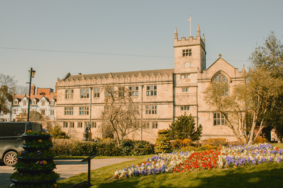A bright, fresh floral arrangement held by a person in front of a historic stone building with a clock tower, featuring a vivid mix of red, purple, and white flowers including tulips and daffodils, wr