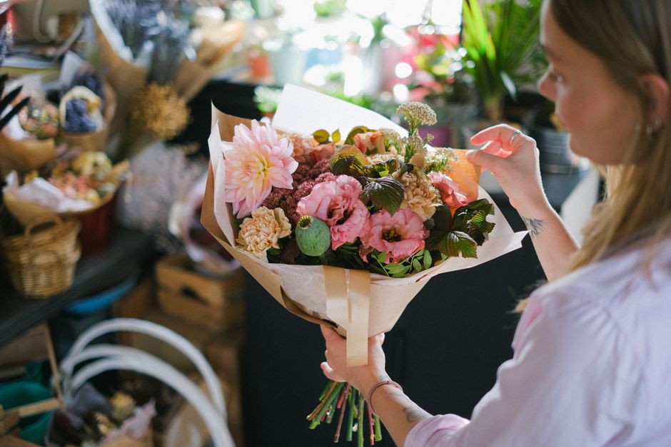 A woman holding a fresh floral bouquet featuring pink and cream flowers, including dahlias, roses, and lisianthus, wrapped in beige and white paper with a satin ribbon. The bouquet is displayed promin
