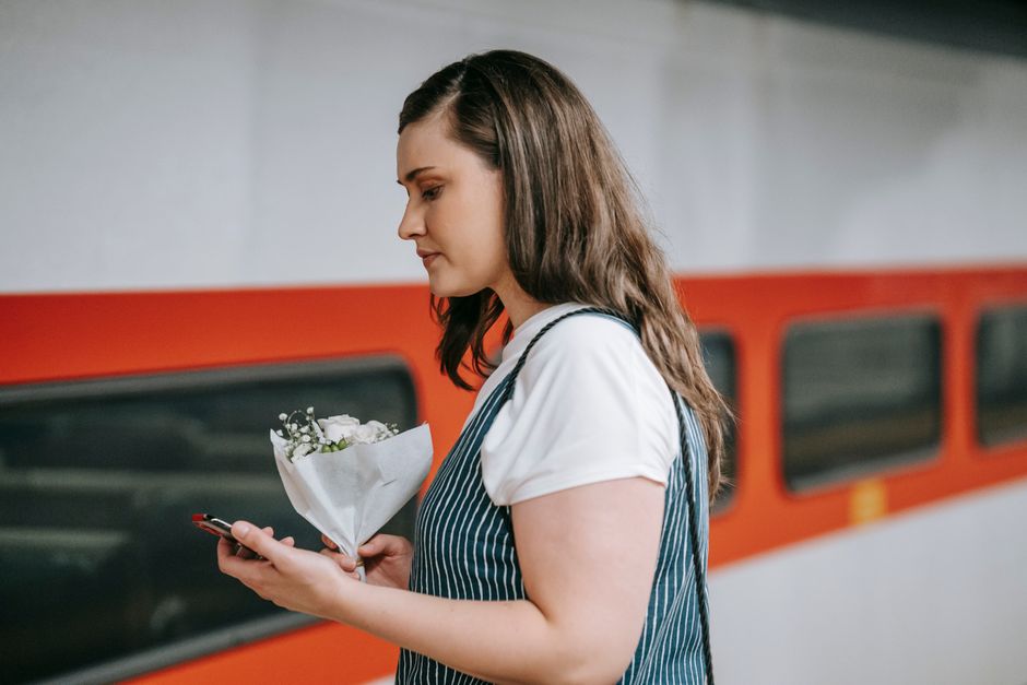 A woman with long, dark brown hair is standing on a train platform, holding a small bouquet of fresh flowers wrapped in white paper. The bouquet features white blooms and green foliage, arranged in a