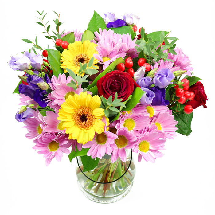 In this image, a radiant mixed bouquet rises from a simple clear glass vase, set against an uncluttered white background that highlights every detail. Deep red roses, their petals plush and tightly spiralled, form the emotional core of the design, offering a feeling of devotion and warmth. Nearby, two sunny yellow gerbera daisies beam with dark brown-orange centres, their broad, flat faces adding an almost cheerful, daisy-like simplicity. Encircling these focal flowers, clusters of pale pink daisies or chrysanthemums fan out with numerous slender petals and tiny yellow centres, building a cushion of softness that gives the bouquet a generous, full shape. Woven between the pink blooms are stems of purple and lavender lisianthus, their gently flared, bell-shaped flowers adding movement and a cool, calming tone that keeps the arrangement from feeling too intense. Little sprays of bright red hypericum berries appear here and there like tiny, lacquered marbles, introducing a tactile contrast to the layered petals. A rich backdrop of mixed greenery holds everything together: broad, glossy leaves support the larger flowers, while finer, lighter green sprigs peek through to add lightness and a sense of natural growth. Through the transparent vase, the viewer can see neatly cut stems arranged with precision in clear water, capturing the craftsmanship you might expect from a local Hertford florist preparing a special order near Fore Street. The bouquet feels joyful yet composed, perfect for marking milestone birthdays in homes around Horns Mill, offering thanks to a teacher at a school near the town centre, or sitting proudly as a vibrant table piece for a family gathering close to Hertford North station.